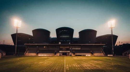 Un stade de football modeste illuminé la nuit, en premier plan, face à l'ombre imposante de trois grandes tours de stades européens à l'horizon, symbolisant le défi d'un club moyen en Ligue des Champions.