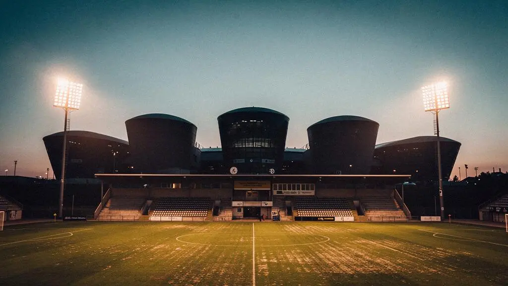 Un stade de football modeste illuminé la nuit, en premier plan, face à l'ombre imposante de trois grandes tours de stades européens à l'horizon, symbolisant le défi d'un club moyen en Ligue des Champions.